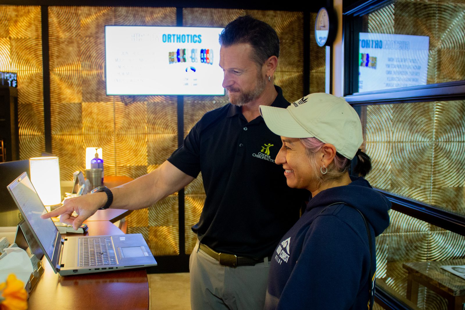 A Cooper Chiropractic provider pointing to a laptop screen while talking with a smiling adult at the clinic’s front desk.