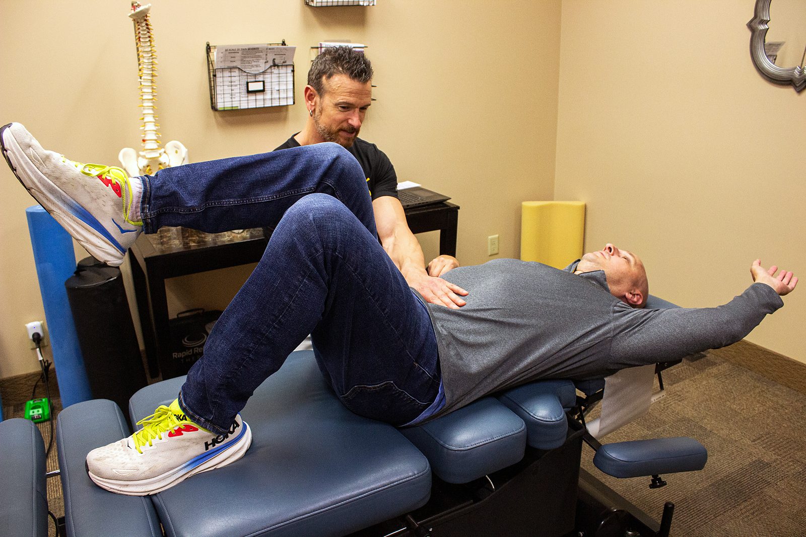 A chiropractor guiding an adult through a dead bug core exercise on a treatment table.